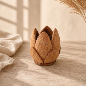 Puede incluir: Un portavelas artesanal de cáscara de coco marrón con forma de flor de loto. El soporte tiene una superficie natural y texturizada y está diseñado para una vela pequeña. El fondo es de color beige claro.