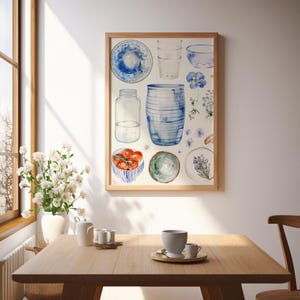 May include: A framed watercolor print featuring various kitchenware items in shades of blue and white. The print includes plates, glasses, a jar, a vase, and bowls with tomatoes and lavender. The artwork is displayed above a wooden table.