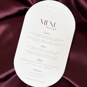 Może przedstawiać: Owalna karta menu z kwiatowym wzorem na górze, prezentująca menu w stylu współdzielonym. Menu zawiera przystawki, dania główne, dodatki i desery. Tekst jest w kolorze ciemnobordowym.