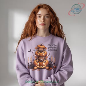 May include: A light purple sweatshirt featuring a stacked pumpkin design with a cow's face and the text "One of thes pumpkins just blinked." The design includes candles, a black butterfly, and roses, perfect for Halloween.