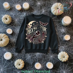 Puede incluir: Sudadera negra con un diseño de Halloween. El diseño presenta una luna creciente, un murciélago, una mano sosteniendo una bola de cristal y el texto "Hexing Hour". La sudadera está rodeada de calabazas blancas y velas.