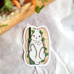 May include: A sticker featuring a white cartoon rabbit inside a sandwich with vegetables. The sticker has a white border and the text "@hellenduu" in the upper right corner. The sticker is on a white surface.