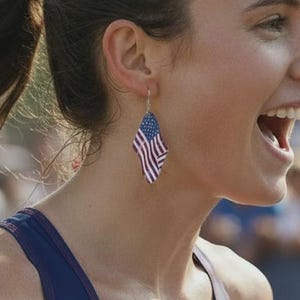 May include: A pair of diamond-shaped dangle earrings featuring the American flag pattern, consisting of a blue field with white stars and red and white stripes. The earrings are worn by a runner in a matching flag-patterned tank top with the text INIMA.