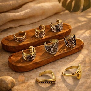 Könnte beinhalten: Eine Sammlung von sieben goldfarbenen Metallringen und Ohrklemmen mit verschiedenen Texturen und Mustern, darunter perlenartige, geometrische und durchbrochene Designs. Die Schmuckstücke sind auf zwei glatten, ovalen Holzplattformen angeordnet.
