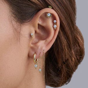 Op de afbeelding: Een collectie goudkleurige oorbellen met heldere ronde stenen. De set bevat een kleine ringoorbel met een hangende steen, een kettingoorbel met een steen en meerdere oorknopjes met heldere stenen langs het kraakbeen en de oorlel.