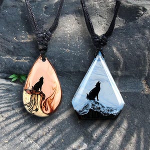 Op de afbeelding: Twee handgemaakte hangerkettingen met zwarte koorden. Eén is druppelvormig met een silhouet van een huilende wolf op een bruine en crèmekleurige achtergrond. De andere is een driehoekige hanger met een wolfsilhouet op een blauw-witte achtergrond.