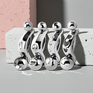 Op de afbeelding: Vier zilverkleurige lichaamsjuwelen. Elk stuk heeft een gebogen ontwerp met een kleine bal aan elk uiteinde en een decoratief element in het midden. Sommige hebben heldere kristallen accenten, terwijl één een hartvormige uitsparing heeft.