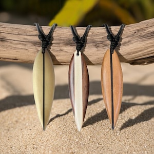 Puede incluir: Tres collares con colgantes de tabla de surf de madera con cordones negros. Los colgantes son en tonos beige claro, marrón y marrón oscuro, cada uno con una raya vertical en el centro. Los collares se exhiben sobre un trozo de madera flotante, con un fondo arenoso.
