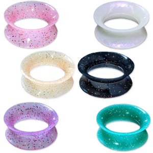 Op de afbeelding: Een verzameling van zes kleurrijke oor stretchers in verschillende tinten, waaronder roze, wit, beige, zwart, paars en teal. Elke stretcher is versierd met glitter, wat een sprankelend effect creëert. De stretchers zijn ontworpen met een uitlopende vorm.