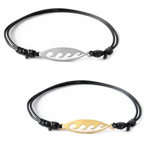 Op de afbeelding: Twee armbanden met zwarte koordbanden en verstelbare knopen. Eén armband heeft een zilverkleurig metalen middendeel in de vorm van een langwerpige ovaal met een uitgesneden golfpatroon. De tweede armband heeft een identiek goudkleurig middendeel.