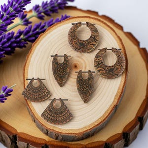Könnte beinhalten: Eine Sammlung von Holzohrringen mit aufwendigen, dunkelbraunen, geätzten Designs. Die Ohrringe haben verschiedene Formen, darunter Creolen und Tränen, und werden auf einer hellen Holzscheibe präsentiert. Lila Lavendelzweige sind im Hintergrund.