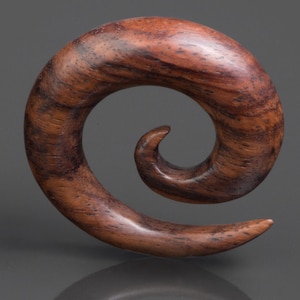 Può includere: Un dilatatore a spirale in legno, di colore marrone scuro con venature più scure. La superficie lucida riflette la luce, mettendo in risalto le venature naturali del legno. Questo pezzo unico è progettato per piercing all'orecchio allargati.