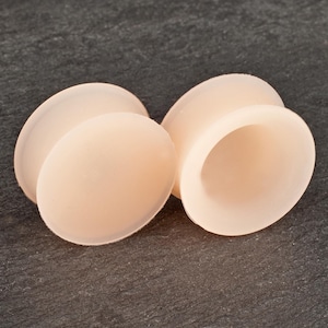 Könnte beinhalten: Zwei hellpfirsichfarbene, oval geformte Ohrstöpsel. Die Stöpsel haben eine glatte, matte Oberfläche und ein ausgestelltes, abgerundetes Design. Die Stöpsel bestehen aus einem weichen Material und sind so konzipiert, dass sie bequem im Ohr sitzen.