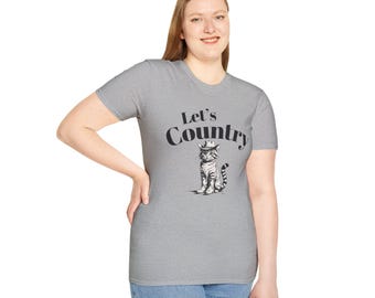 Op de afbeelding: Een grijze t-shirt met de tekst "Let's Country" en een afbeelding van een kat met een cowboyhoed en -laarzen. De kat zit. Het t-shirt heeft korte mouwen.