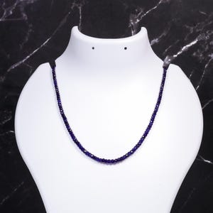 Peut inclure: Un collier délicat composé de petites perles bleu foncé. Le collier est présenté sur un buste de mannequin blanc, sur un fond sombre et marbré. Les perles semblent être de taille uniforme et étroitement enfilées.