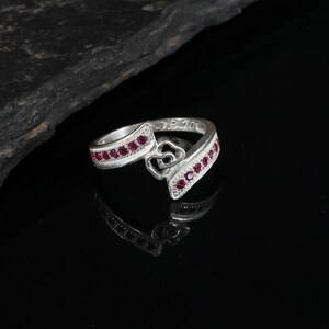 May include: A silver-toned ring featuring a bypass design with two interlocking heart shapes at the centre. The band is accented with a row of small, round, deep pink stones embedded along each side of the split shank, creating a symmetrical look.