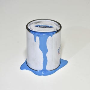 May include: A white paint tin with a blue lid and the word "Ford" printed on it, with blue paint spilling out. A small American flag is visible on the side. The paint is pooling around the base.