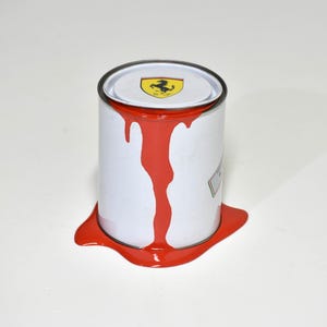 May include: A white paint tin with red paint overflowing. The lid features a black and yellow Ferrari logo. Red paint drips down the side and pools around the base of the tin.