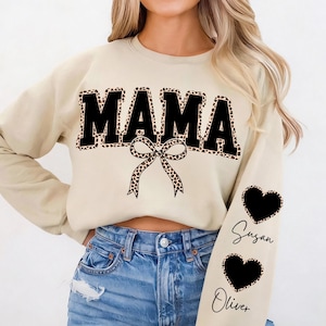 Puede incluir: Sudadera beige con la palabra "MAMA" en letras negras grandes con borde de estampado de leopardo. Un lazo con estampado de leopardo está debajo de la palabra. La manga izquierda tiene dos corazones negros con estampado de leopardo con los nombres "Susan" y "Oliver".