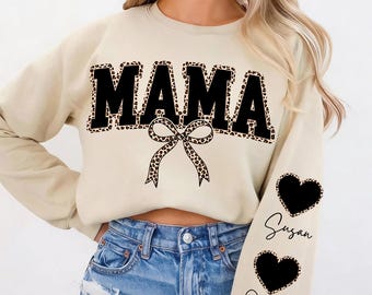 Puede incluir: Sudadera beige con la palabra "MAMA" en letras negras grandes con borde de estampado de leopardo. Un lazo con estampado de leopardo está debajo de la palabra. La manga izquierda tiene dos corazones negros con estampado de leopardo con los nombres "Susan" y "Oliver".