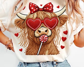 Pode incluir: Camiseta creme com bordado de vaca Highland com óculos de sol em forma de coração e laço vermelho. A vaca está segurando um pirulito. Corações vermelhos estão espalhados ao redor do desenho. Um design divertido e extravagante.