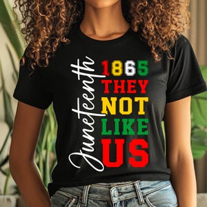 May include: Black t-shirt with the word "Juneteenth" in white script on the left side. The shirt features the text "1865 THEY NOT LIKE US" in red, yellow, and green block letters.