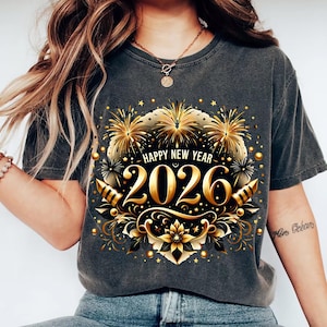 May include: Dark gray t-shirt with a gold and black graphic design for the New Year 2026. The design includes fireworks, floral elements, and the text "HAPPY NEW YEAR". The shirt is a casual style.