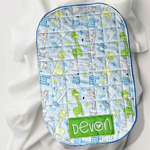 May include: A baby changing mat with a light blue quilted surface featuring a giraffe pattern in various colours. It has a blue border and a green patch with the name "Devon" embroidered on it.