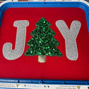 May include: Red fabric with the word "JAY" in silver glitter letters and a green sequined Christmas tree. The design is set in a blue embroidery hoop. The text "Keep Away From Computer Disks and Hard Drives" is visible.