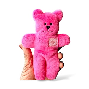 May include: A bright pink plush teddy bear with a stitched black nose and mouth. The bear has a small square patch on its chest. The bear is held up against a white background.