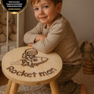 Op de afbeelding: Een houten kruk met een Rocket Man-ontwerp. De ronde bovenkant van de kruk toont een astronaut op een raket, met de woorden "Rocket man" eronder gegraveerd. Hij heeft drie houten poten. De afbeelding bevat ook een kind en een prijs.