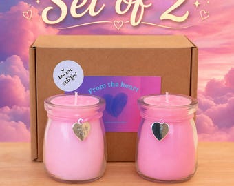Pode incluir: Um conjunto de duas velas cor-de-rosa em frascos de vidro transparente, cada uma com um pendente prateado em forma de coração atado ao pescoço. As velas estão em frente a uma caixa de cartão castanha com a etiqueta From the heart e Set of 2.