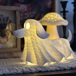 May include: A decorative, illuminated dog-shaped lamp with a flowing, draped design. The lamp emits a warm glow, with the eyes and a mushroom-shaped lamp in the background lit up. The lamp is white and sits on a textured surface.