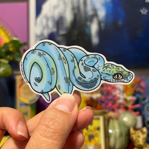 May include: A sticker depicting a blue snake with green and black patterns. The snake is coiled, with a detailed head and eye. The sticker has a white border and the artist's name is printed on the left side. A decorative item.