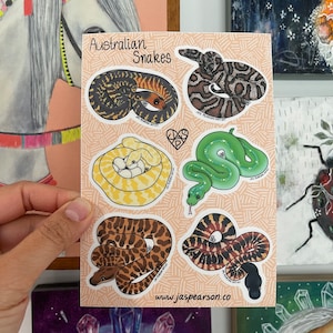 Peut inclure: Un ensemble de 6 autocollants de serpents australiens sur un fond rose à pois blancs. Les autocollants présentent différentes espèces de serpents, dont un serpent arboricole vert, un serpent brun et un serpent jaune. Les autocollants sont tous illustrés dans un style cartoon et ont une bordure blanche. Le texte "Australian Snakes" est en haut de la feuille. Le texte "www.jaspearson.co" est en bas de la feuille.