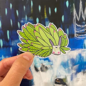 Może przedstawiać: Naklejka przedstawiająca kreskówkową ślimak morską z zielonym korpusem i różowymi końcówkami. Ślimak ma fioletowe uszy i różową twarz z czarnymi oczami. Naklejka znajduje się na białym tle.