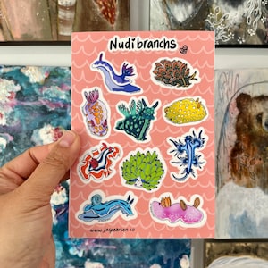 Może przedstawiać: Arkusz naklejek z kolorowymi ilustracjami ślimaków nagoskrzelnych, znanych również jako ślimaki morskie. Naklejki znajdują się na różowym tle z falistym wzorem. Tekst "Nudibranchs" znajduje się u góry arkusza. Adres strony internetowej "www.jaspearson.co" znajduje się u dołu arkusza.