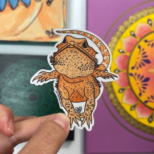 May include: A sticker of an orange and tan bearded dragon with its arms spread. The sticker has a white border and the artist's signature. The lizard has a textured appearance and a heart symbol near its feet.
