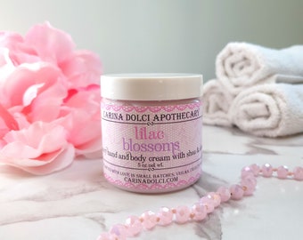 Könnte beinhalten: Ein Tiegel "Lilac Blossoms" Hand- und Körpercreme mit Sheabutter. Das Etikett ist helllila mit weißem Text und einem floralen Design. Der Tiegel hat 142g Nettogewicht und ist von rosa Blumen, einer Perlenkette und gerollten Handtüchern umgeben.