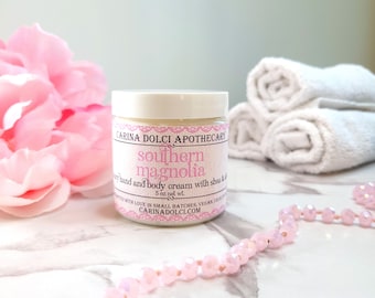 Könnte beinhalten: Ein weißes Glas mit einem rosa Etikett, auf dem "CARINA DOLCI APOTHECARY southern magnolia hand and body cream with shea & 5 oz net wt. WITH LOVE IN SMALL BATCHES, VEGAN, CRUELTY-FREE CARINADOLCI.COM" steht.