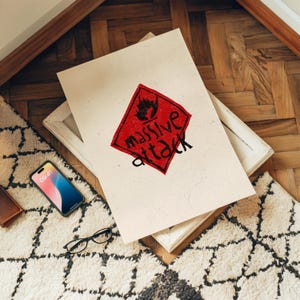 Puede incluir: Una impresión enmarcada con las palabras "massive attack" en negro sobre un rombo rojo. La impresión descansa sobre un marco blanco, con un teléfono inteligente, gafas y una cartera cerca. El fondo incluye una alfombra estampada y un suelo de madera.