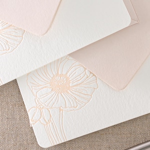 May include: Close-up of stationery featuring cream cards and blush envelopes. The cards have an embossed floral design in a soft peach colour. The envelopes are a solid blush colour, creating a delicate and elegant aesthetic.