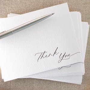 May include: A pile of white 'Thank You' cards with black calligraphy, and a silver pen resting on top. The cards are on a textured surface, with vintage postage stamps visible in the background.