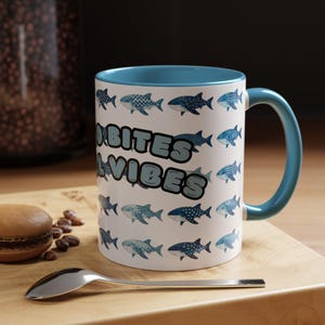 May include: White ceramic mug with a blue interior and handle, decorated with various fish illustrations and the text "Good Vibes". A spoon and a macaron are on a wooden surface.