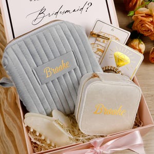 May include: A gift box with the text "WILL YOU BE MY Bridesmaid?" contains a gray velvet cosmetic bag, a white jewelry case, hair accessories, and a diamond-shaped card. The name "Brooke" is embroidered on the bags. A pink ribbon and flowers complete the presentation.