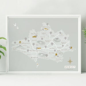 May include: A gray and gold map print of Dorset, England, with the names of towns and cities labeled in brown text. The map is framed in a white frame and is hanging on a white wall.