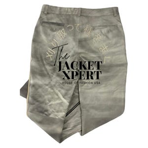 May include: Light gray pants with a subtle gradient effect. The back of the pants features Japanese text embroidery and the text "The Jacket Xpert, House of Fashion USA". The pants have a zipper detail and a vertical stripe.