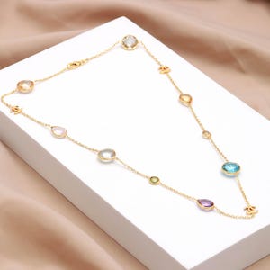 May include: A gold necklace with a fine chain, adorned with a variety of colourful gemstones. The necklace showcases oval, round, and teardrop-shaped stones in blue, purple, yellow, and green. The necklace is presented on a white surface.
