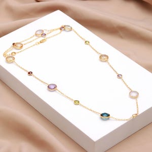 May include: A gold-colored necklace with various colorful gemstones. The necklace features a delicate chain with multiple round and oval-shaped stones in shades of purple, blue, yellow, and red. The necklace is displayed on a white surface.