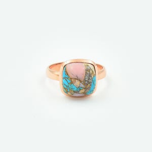 May include: A rose gold ring with a square, multi-colored stone. The stone features a mix of pink, turquoise, and gold hues. The ring has a simple band and a bezel setting, showcasing the unique stone.
